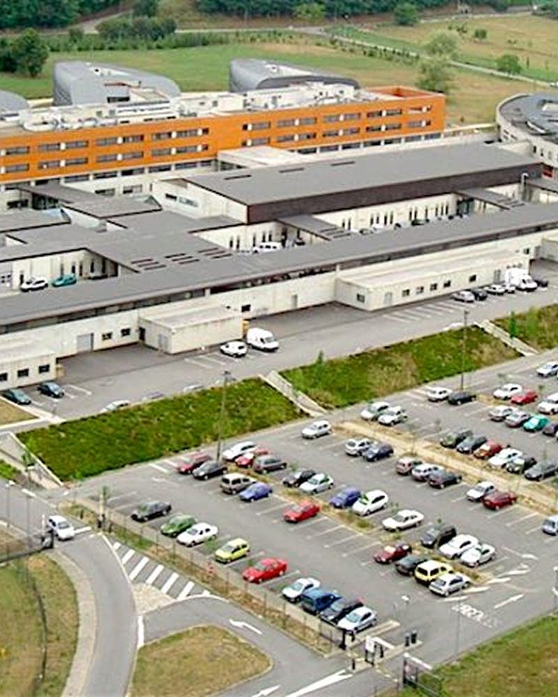 An aerial view of a hospital campus, its orange and grey wings set above a full car park and open countryside