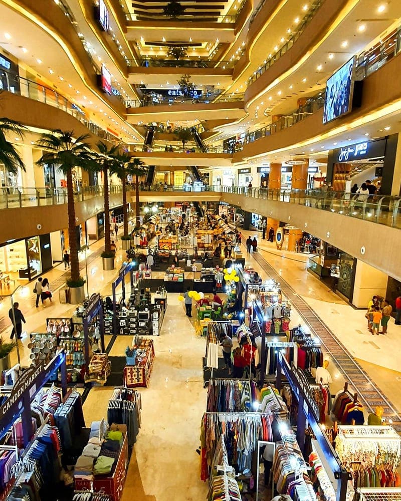 The bright multi-level atrium of a shopping centre, palms and market stalls filling the floor below